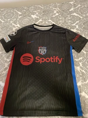 Svarta Nike FC Barcelona tröja - Säljer en svart FC Barcelona matchtröja från Nike med röda och blå detaljer på sidorna. Tröjan har Spotify-logga på bröstet, broderat FCB-märke och Lamine Yamal 19 på ryggen. Materialet är lätt och andas, perfekt för fotboll eller träning.