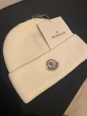 Vit Moncler stickad mössa - Säljer en snygg vit stickad mössa från Moncler med klassisk Moncler-logga framtill. Mössan är ribbstickad och har en bred uppvikt kant. Perfekt för dig som vill ha en stilren och lyxig accessoar till vintern.