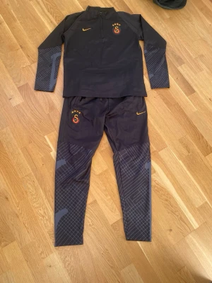 Galatasaray x Nike svart träningsset - Svart träningsset från Nike och Galatasaray med långärmad tröja och matchande byxor. Setet har orange Nike-logga och Galatasaray-emblem på både tröja och byxa. Ärmar och benslut har grått grafiskt mönster. Materialet är syntet och tröjan har half zip.