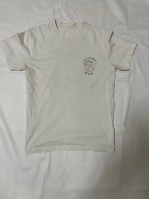 Vit Casablanca Airways t-shirt - Säljer en vit t-shirt från Casablanca med tryck fram och stort grafiskt tryck på ryggen med texten 'Casablanca Airways' och motiv av ett flygplan, blad och blomma. T-shirten har rund hals och korta ärmar, perfekt för chill dagar.