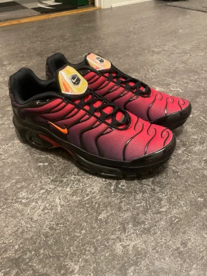 Nike Air Max Plus Tn röd/svart - Nike Air Max Plus Tn sneakers med grymt röd och svart färgskiftning, orange swoosh och coola svarta detaljer. Ovandelen har vågiga linjer och meshmaterial, medan sulan har synliga Air-enheter och färgglada inslag undertill. Perfekt för dig som vill sticka ut med dina kicks.
