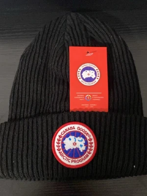 Svart ribbad mössa  - Svart ribbstickad mössa med klassisk rund patch framtill i rött, vitt och blått. Mössan har uppvikt kant och är tillverkad i ett mjukt stickat material som håller värmen. Perfekt för kalla dagar och snygg till streetwear.