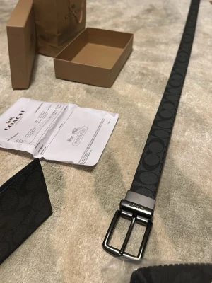 Coach svart mönstrad bälte - Snyggt svart bälte från Coach med deras ikoniska logomönster i mörkgrått. Bältet har en stilren metallspänne med Coach-gravyr och är tillverkat i slitstarkt syntetmaterial med skinnkänsla. Perfekt för att lyfta din outfit med en lyxig touch.