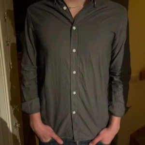 Grå skjorta från These Glory Days - Säljer en stilren grå skjorta från These Glory Days i storlek S. Skjortan har klassisk krage, vita knappar framtill och långa ärmar. Perfekt för dig som gillar enkel och clean look. Materialet känns mjukt och lätt mot huden.