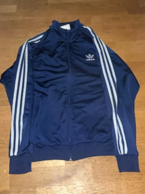 Blå Adidas vindjacka med ränder - Snygg marinblå vindjacka från Adidas med klassiska vita ränder längs ärmarna och logga på bröstet. Jackan har dragkedja framtill och ribbade muddar vid ärmslut och nederkant. Perfekt för en sportig och chill stil.