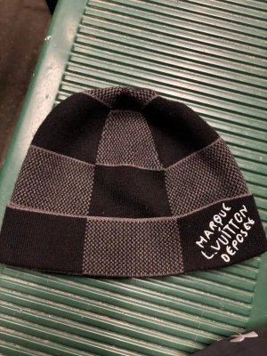 Rutig svart och grå Louis Vuitton mössa - Säljer en snygg mössa från Louis Vuitton i svart och grått med rutigt mönster. Mössan har broderad text 'Marque L. Vuitton déposée' på kanten och är stickad i mjukt material. Perfekt för dig som vill ha en stilren accessoar med lyxig känsla.