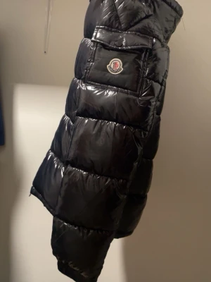 Svart pufferjacka från Moncler - Säljer en svart pufferjacka från Moncler med glansig finish och klassisk logga på ärmen. Jackan har dragkedja framtill, quiltad design och innerficka med serietidningsdetalj. Perfekt för kalla dagar och har en riktigt snygg streetstil.