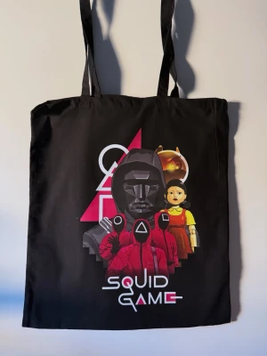Svart Squid Game shoppingväska - Cool svart tygkasse med tryck från Squid Game. Väskan har långa handtag och motiv med karaktärer och symboler från serien i rött, gult och rosa. Perfekt för att bära böcker, laptop eller shopping. Snygg accessoar för fans av serien!