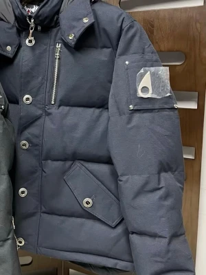 Moose Knuckles mörkblå pufferjacka - Säljer en mörkblå pufferjacka från Moose Knuckles med stor huva och fluffig pälsdetalj. Jackan har silverfärgade knappar, dragkedja, och en ikonisk logga på ärmen. Flera fickor med knappstängning och quiltad design ger en cool och varm look.