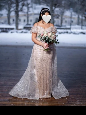 Glittrig champagnefärgad festklänning - Säljer en magisk långklänning i champagnefärg med glittriga paljetter över hela klänningen. Offshoulder-modell med puffiga ärmar och v-ringad urringning. Klänningen har en elegant släp och är figurnära i modellen. Perfekt för bal eller fester!