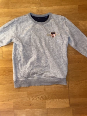 Grå sweatshirt från GANT - Snygg grå sweatshirt från GANT med broderad logga och emblem på bröstet. Tröjan har rund hals, ribbade muddar och är långärmad. Perfekt för dig som gillar klassisk och sportig stil. Passar dig som är ca 170 cm lång.