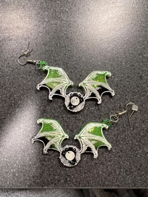 Grön svarta fladdermus örhängen - Cool örhängen med gröna och svarta nyanser, formad som fladdermusvingar. Örhängena har vita detaljer av skelett fladdermusvingar och ett öga i mitten. Perfekt för dig som vill sticka ut med en unik look och edgy vibe.