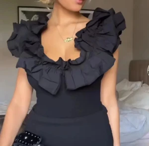 Svart body med volangdetaljer - Supersnygg svart body med djup v-ringning och stora volanger längs axlar och urringning. Bodyn har öppen rygg och en stilren, elegant look som verkligen passar till fest eller vardag. 