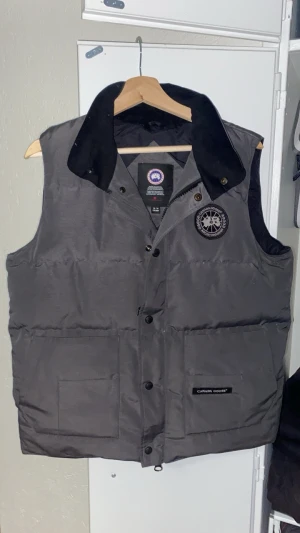 Mörkgrå dunväst från Canada Goose - Hej, säljer en mörkgrå dunväst från Canada Goose med hög krage, stora framfickor och diskret logga på bröstet. Osäker på om den är äkta men allting ser perfekt ut på den så jag är väldigt säker på att den är äkta. Pris går att diskutera.