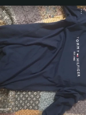 Mörkblå långärmad topp Tommy Hilfiger - Säljer en mörkblå långärmad topp från Tommy Hilfiger med tryckt logga och texten 'EST. 1985' på bröstet. Toppen har en klassisk och stilren design, perfekt för dig som gillar ikoniska märken och enkel stil.
