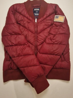 Mörkröd pufferjacka från Svea - Säljer en snygg mörkröd pufferjacka från Svea med ribbade muddar och krage. Jackan har ett broderat Svea-märke på bröstet och en patch på ärmen. Dragkedja framtill och quiltad design som ger extra volym och värme.