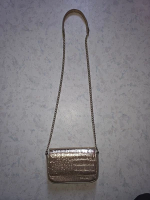 Guldig axelväska från H&M - Säljer en snygg guldig axelväska från H&M med krokodilpräglad yta och kedjerem. Väskan har ett magnetlås och ett innerfack. Perfekt storlek för det viktigaste och ger en lyxig touch till din outfit.