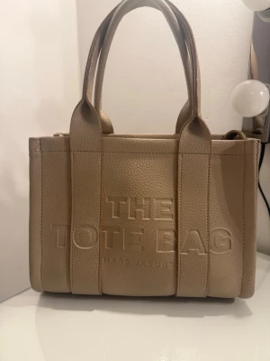 Beige Tote Bag  - Säljer en beige Tote Bag i äkta skinn med präglad logga framtill. Väskan har två rejäla handtag, strukturerad form och dragkedja upptill. Insidan är rymlig med svart foder och en innerficka. ❤️