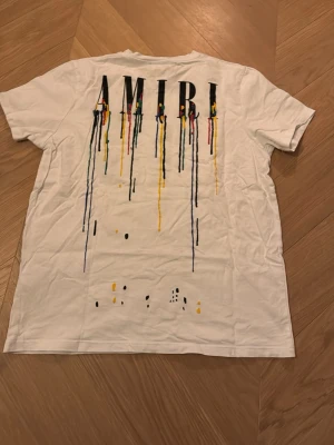 Vit AMIRI t-shirt med färgstänk - Säljer en vit t-shirt från AMIRI med coolt tryck på både fram- och baksidan. Stora AMIRI-bokstäver med färgstänk i svart, gul, röd, grön och blå som rinner ner över tyget. Klassisk rund hals och korta ärmar. Materialet är mjuk bomull, perfekt för en chill och trendig look.