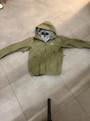 Grön vindjacka från Arc'teryx - Arc'teryx vindjacka i olivgrön färg med huva och hel dragkedja framtill. Jackan har två sidofickor med dragkedja och justerbar nederkant. Tillverkad i lätt syntetmaterial, perfekt att packa ihop och ta med. Snygg logga på bröstet och minimalistisk design.