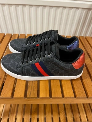 Gucci ace skor - Säljer ett par svarta Gucci ace skor med GG-mönster, vita platta sulor och snörning. Sidorna har röd och marinblå rand, och hälen har metallicröd och metallicblå ormskinnsdetalj med Gucci-logga. Materialet är skinn och syntet, och tån är rund. Storlek 43 mycket bra skick