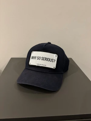 Marinblå John Hatter & Co keps  - Snygg marinblå keps från John Hatter & Co med mesh på sidorna och bak. Framtill sitter en metallskylt med texten 'WHY SO SERIOUS?'. Kepsen har böjd skärm och justerbar passform. Perfekt för dig som vill sticka ut med en cool detalj. Kepsen är i väldigt bra skick och priset går att diskuteras. Det är bara att höra av sig om man har någon fråga.
