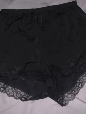 Svarta shorts med spetsdetaljer - Säljer ett par svarta shorts med bred resår i midjan och snygga spetsdetaljer längs bensluten. De har en mjuk och glansig yta som ger en lyxig känsla, perfekta för att chilla hemma eller styla upp med en cool topp.
