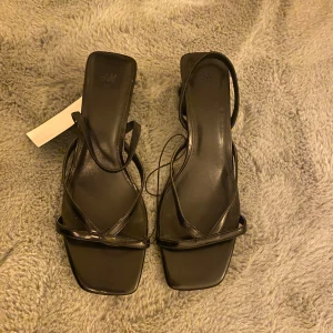 Svarta klackar från H&M - Snygga svarta sandaler från H&M med fyrkantig tå och smala remmar. Skorna har en låg klack och är tillverkade i syntetmaterial med stilren design. Perfekta för dig som gillar minimalistisk och trendig look.