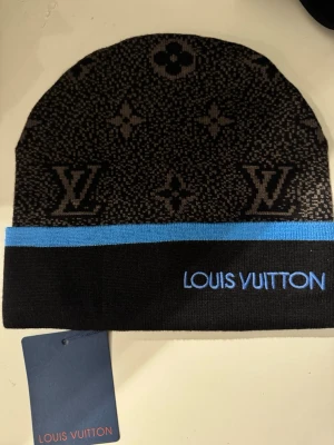 L-V stickad mössa med blå kant - Snygg stickad mössa från L-V i svart och grått med klassiskt L-V-monogram och blommönster. Mössan har en blå kant och L-V-broderi framtill. Perfekt för dig som vill ha en lyxig accessoar med ikonisk design.