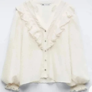Vit volangblus med prickar från Zara - Supersöt vit blus från Zara med volangdetaljer längs v-ringningen och ärmsluten. Prickigt mönster i tyget, knappar framtill och luftiga, långa ärmar. Perfekt för dig som gillar romantisk och trendig stil. Ena armen är lösare än den andra, men inget man märker