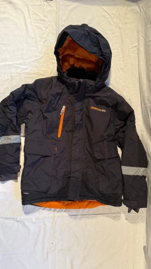 Svart vindjacka från Didriksons 1913 - Svart vindjacka från Didriksons 1913 med orange foder och detaljer. Jackan har huva, flera fickor, reflexdetaljer på ärmar och rygg samt dragkedja framtill. Perfekt för blåsiga dagar och har en cool, sportig vibe med Storm System-teknologi. Pris kan diskuteras via snabb affär 