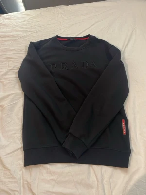 Svart sweatshirt från Prada i bomull - Säljer en svart sweatshirt från Prada med diskret broderad logga framtill och röd detalj i nacken. Tröjan är i 100% bomull, har rund hals och långa ärmar. Perfekt för dig som gillar stilrena designer och exklusiva märken.skulle säga att tröjan passar mellan s-m, bara att höra av er om ni har funderingar 😁