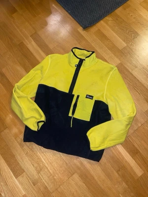 Gul och svart fleece från Penfield L - Säljer en gul och svart fleece från Penfield i storlek L. Tröjan har en halv dragkedja, hög krage och en praktisk bröstficka med dragkedja. Tillverkad i mjuk fleece som är perfekt för kyliga dagar. Snygg färgblockad design och bekväm passform.