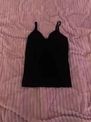 Svart ribbad linnetopp med smala axelband - Säljer en enkel och stilren svart ribbad  linnetopp med smala axelband och rak passform. Toppen är ärmlös och har en klassisk look som passar till många olika outfits. Perfekt att matcha med jeans eller kjol.