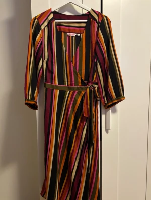 Randig omlottklänning från Indiska - Färgstark omlottklänning från Indiska med vertikala ränder i svart, orange, rosa, olivgrön och beige. Klänningen har trekvartsärm, v-ringning och knyt i midjan. 