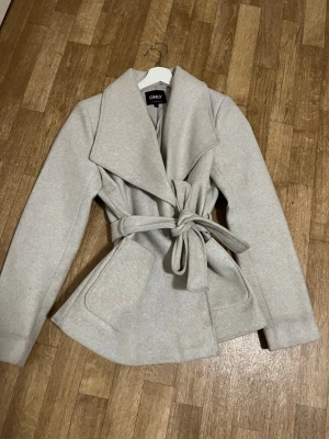 Beige omlott kappa från ONLY - Snygg beige kappa från ONLY med bred krage och knytbälte i midjan. Jackan har stora fickor framtill och en klassisk omlottmodell som ger en clean och stilren look. Perfekt för dig som vill ha en enkel men trendig jacka.