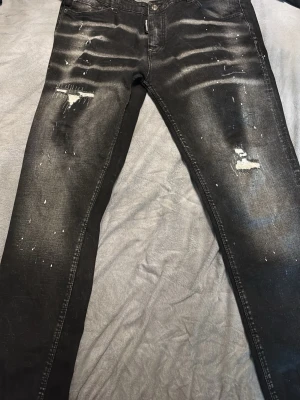 Svarta Dsquared2 jeans med slitningar - Säljer ett par svarta Dsquared2 jeans med coola slitningar, färgstänk och patchade detaljer på framsidan. Jeansen har fem fickor, knappgylf och en snygg tvättad look. Märkespatch bak i midjan och klassisk rak passform. Perfekta för dig som gillar streetwear.