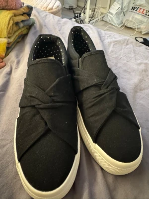 Svarta slip-on sneakers med knutdetalj - Snygga svarta slip-on sneakers från Anna Field med unik knutdetalj över foten. Skorna har vit sula och insida med prickigt mönster. Tillverkade i textilmaterial för en skön och avslappnad look. Perfekta till jeans eller kjol för en chill vardagsstil.