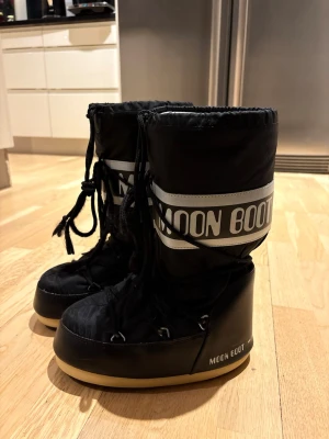 Svarta Moon Boots med snörning - Moon Boots i svart med vita detaljer och tydlig logga runt skaftet. Skorna har snörning framtill och en platt, mönstrad sula i svart och beige. Yttermaterialet är syntet och insidan är fodrad för extra värme. Perfekta för kalla vinterdagar. Varmaste vinterskorna jag testat, är något snetrampade och tyget på insidan är något trasigt, men inget man tänker på, pris går att diskutera, storlek 35-38
