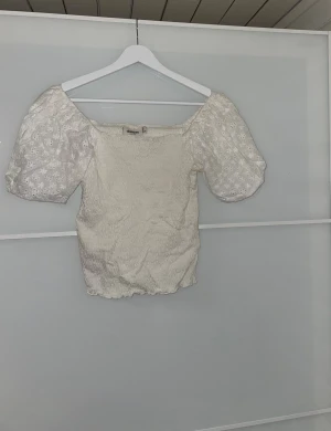 Vit blus med puffärmar från MQ - Supersöt vit blus från Dobber med smockad kropp och broderade puffärmar i blommigt mönster. Blusen är vackert sydd med volangkant nedtill och i 100% Bomull. Perfekt för vår och sommar!