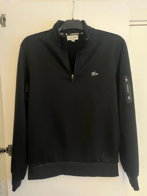 Svart half zip tröja från Lacoste - Säljer en svart långärmad tröja från Lacoste med half zip och klassisk krokodillogo på bröstet. Tröjan har en diskret ficka med dragkedja på ärmen. Pris kan diskuteras vid snabb affär.