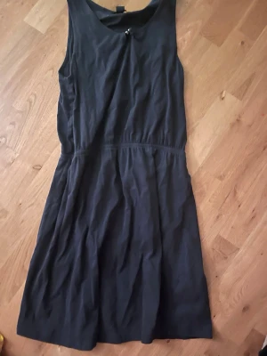 Svart ärmlös klänning från Esmara - Säljer en svart ärmlös klänning från Esmara med en söt detalj i form av knyt och små pärlor vid halsringningen. Klänningen har en markerad midja och är tillverkad i mjuk bomull, perfekt för en enkel och stilren look.