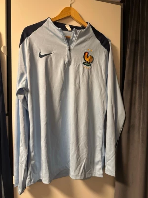 Nike Frankrike fotbollsträningsoverall - Säljer en Nike träningsoverall från franska landslaget med långärmad ljusblå tröja och marinblå byxor. Båda plaggen har FFF-logga och Nike Swoosh. Tröjan har half zip och byxorna har resår i midjan. Perfekt för dig som älskar fotboll och vill ha en clean look.