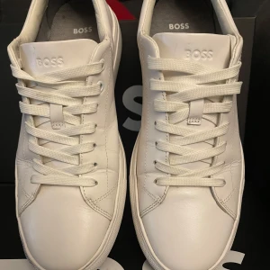 Vita sneakers från BOSS i skinn - Stilrena vita sneakers från BOSS med klassisk rund tå och platt sula. Skorna är tillverkade i slätt skinn och har vita snören samt diskret BOSS-logga på plösen och sulan. Perfekta för en clean och modern look. Använda en gång.