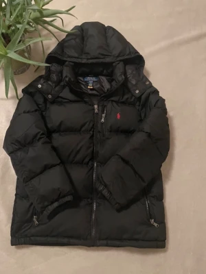 Svart pufferjacka från Polo Ralph Lauren - Säljer en svart pufferjacka från Polo Ralph Lauren med röd broderad logga på bröstet. Jackan har huva, dragkedja framtill och är vadderad för extra värme. Perfekt för kalla dagar och har en clean, sportig look.