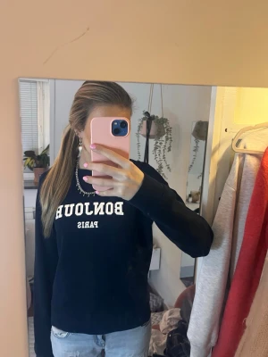 Mörkblå sweatshirt från Holly & Whyte - Mörkblå sweatshirt med trycket 'BONJOUR PARIS' i vitt framtill. Klassisk rund halsringning och långa ärmar. Tillverkad i mjuk bomullsmix, perfekt för en chill och stilren look.