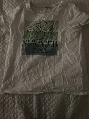 Vit t-shirt med tryck från Street One - Vit t-shirt från Street One med coolt tryck på framsidan där det står 'SUMMER SECRETS' och en stiliserad blomma. T-shirten har rund hals och korta ärmar, perfekt för varma dagar. Materialet är mjukt och känns skönt mot huden.