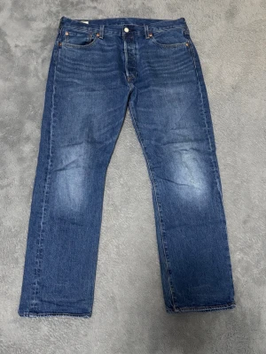 Levi's 501 blå jeans - Klassiska blå Levi's 501 jeans med raka ben och normal passform. Jeansen har fem fickor, knappgylf och den ikoniska läderpatchen bak i midjan. Tillverkade i slitstarkt bomullsjeansmaterial med snygg tvätt och subtila slitningar.