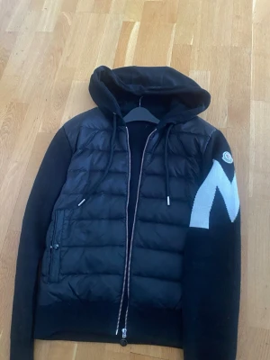 Moncler - Säljer en svart pufferjacka från Moncler med huva och dragkedja. Jackan har stickade ärmar med en stor vit M-detalj på ena ärmen och Moncler-logga. Framsidan är quiltad och har fickor med knappstängning. Perfekt för dig som vill ha en snygg och sportig look. Väldigt sällsynt jacka dessvärre priset 