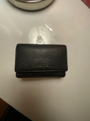 Svart plånbok från Polo Ralph Lauren - Säljer en stilren svart plånbok från Polo Ralph Lauren i äkta skinn. Plånboken har en klassisk design med tryckknapp och präglad logga på framsidan. Perfekt storlek för kort och sedlar, med en enkel och clean look.